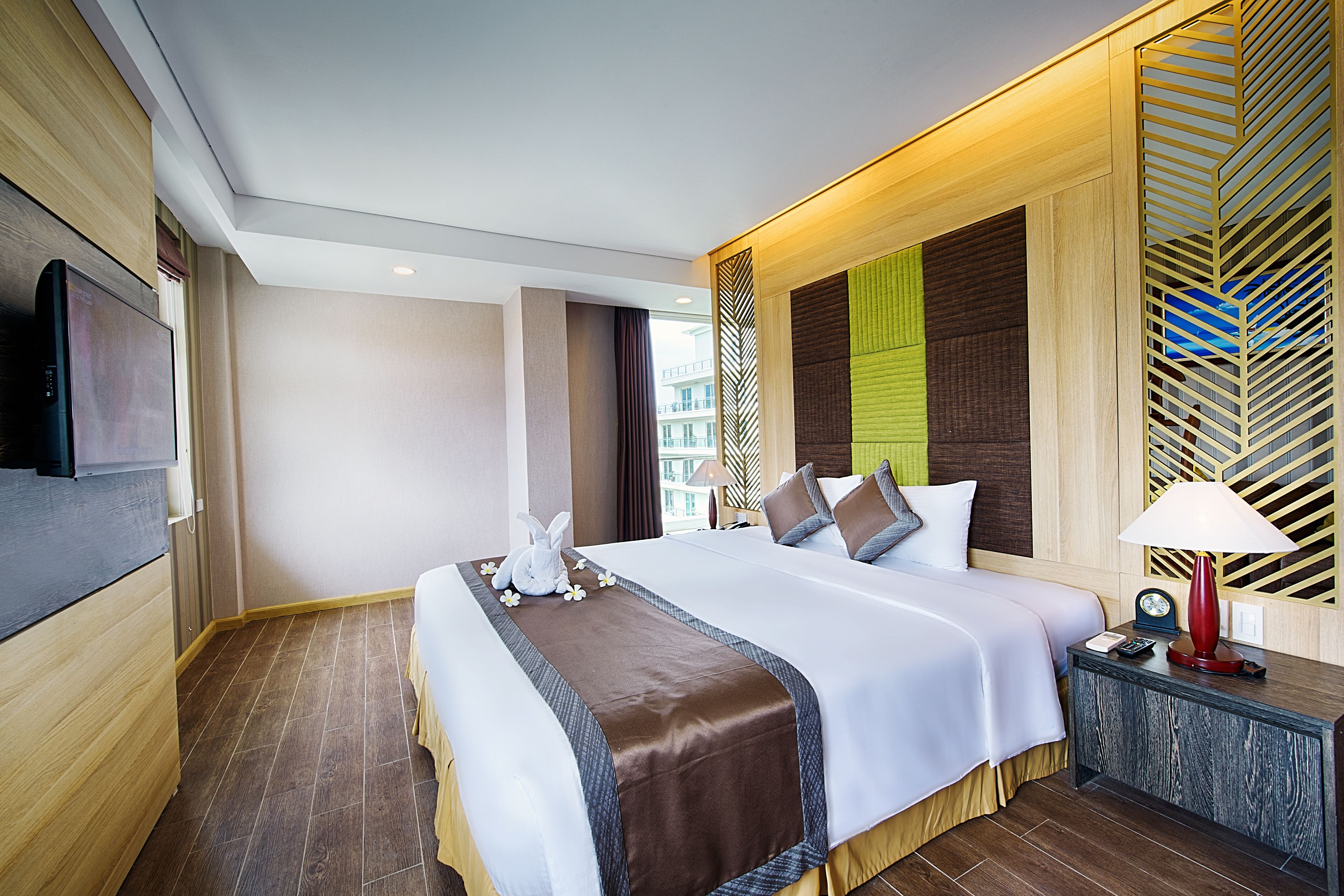 A hotel room with a large bed, a TV, a bedside table with a lamp, and a view of the city.