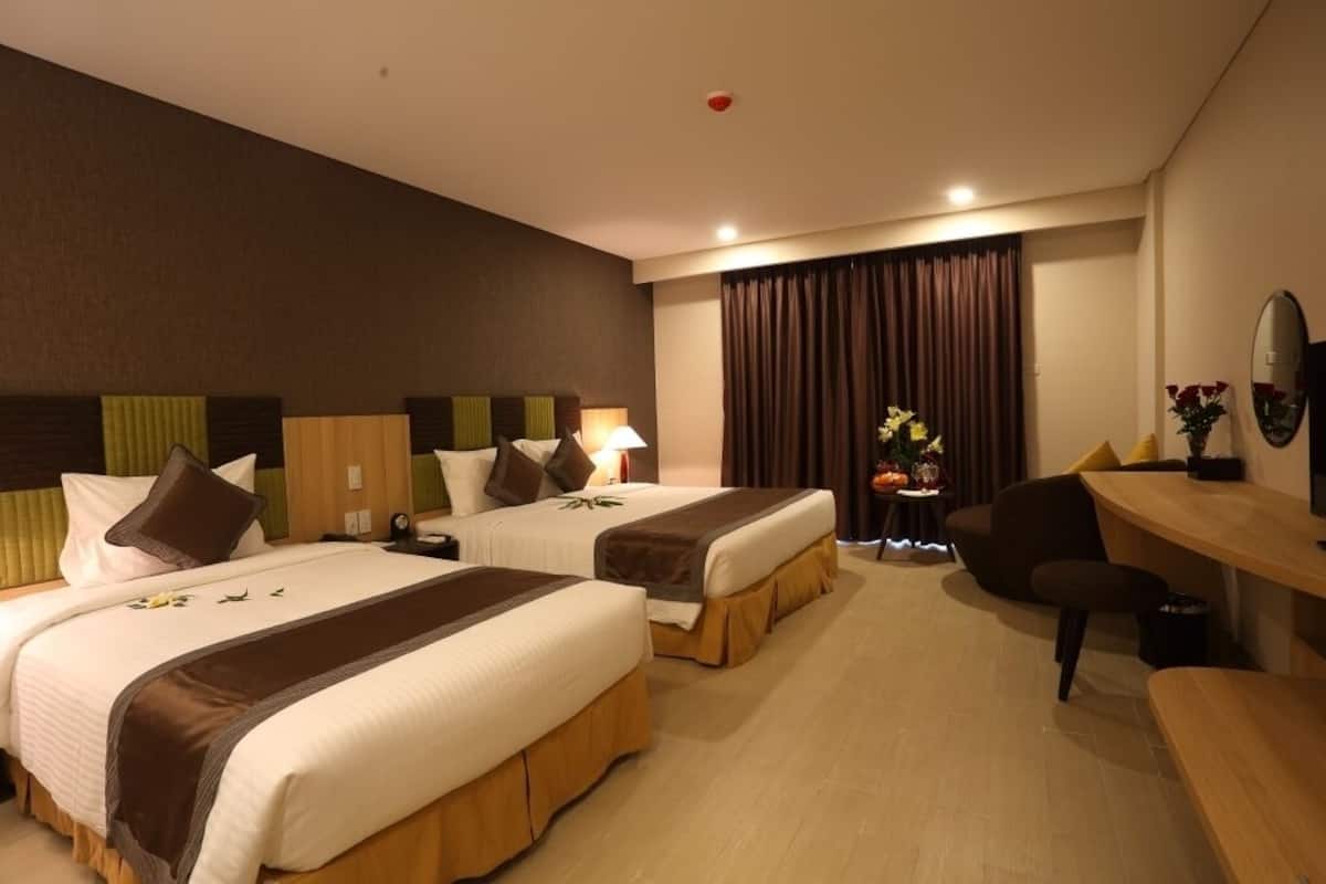 A hotel room with two beds, a desk, a chair, a small table with flowers, and a mirror on the wall.