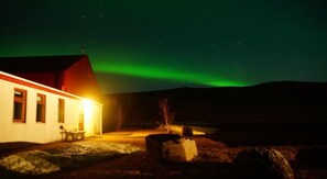 A building with a red roof and a lit interior, set against a backdrop of the northern lights. - Hótel Selið (Rangárþing ytra)
