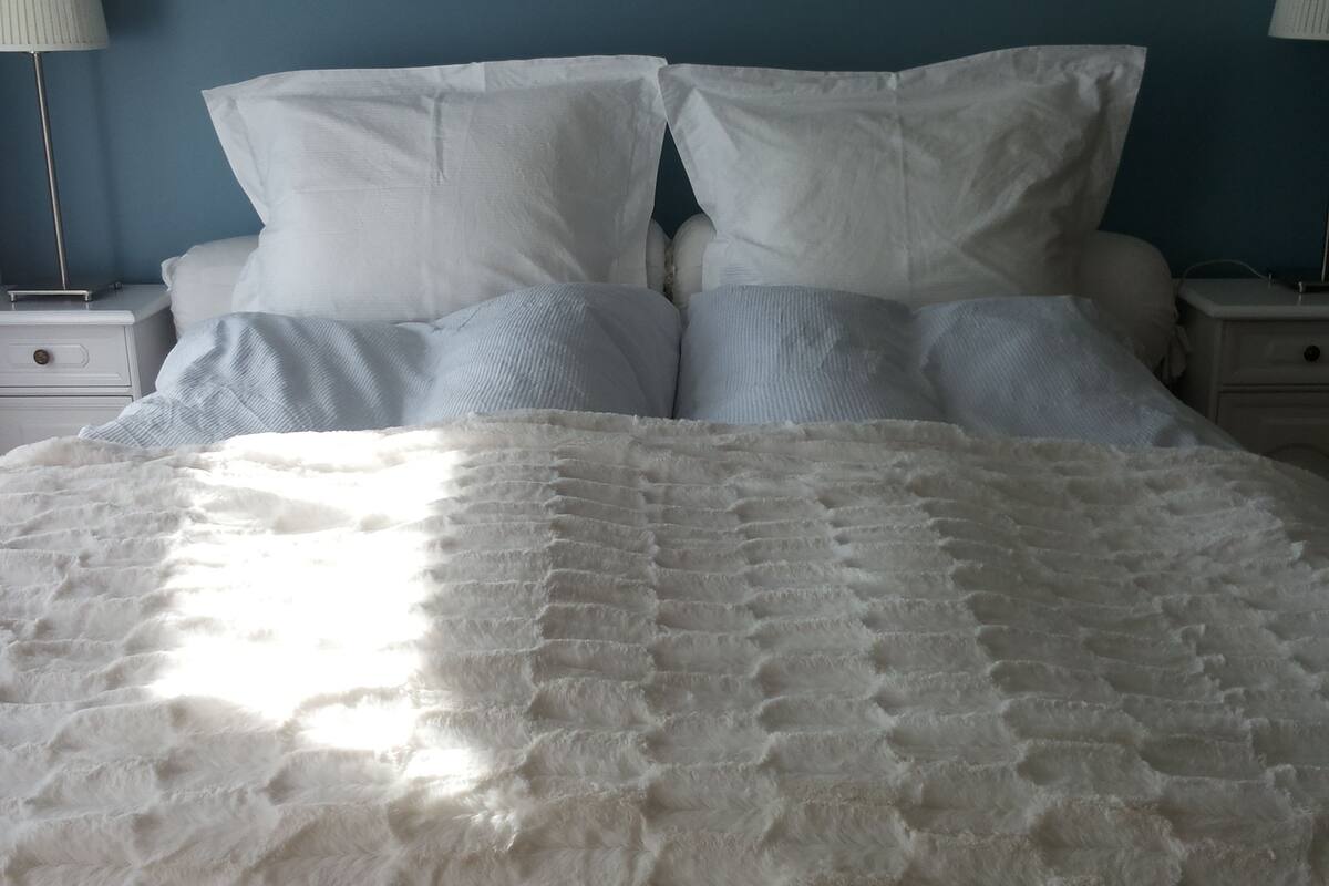 Una cama con un edredón blanco texturizado, dos almohadas blancas y una manta azul claro. Hay dos mesitas de noche con lámparas y un cajón pequeño.