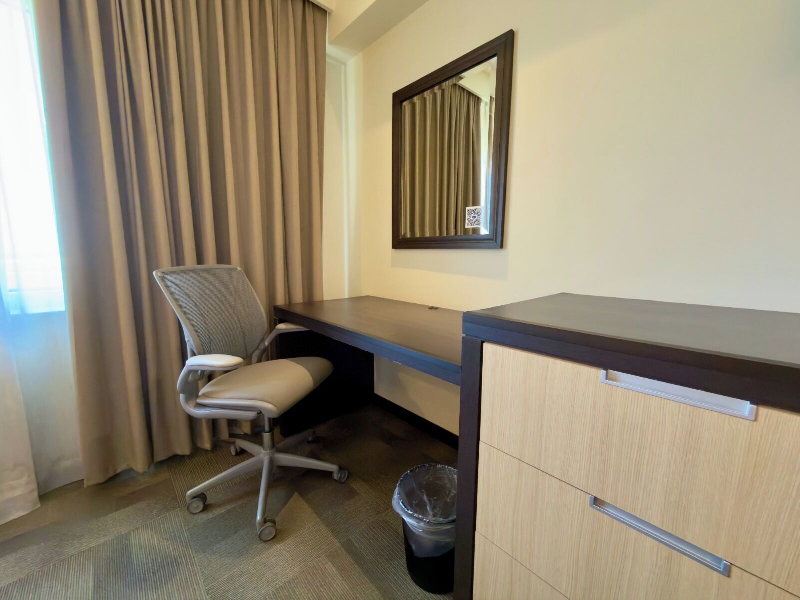 A hotel room with a desk, chair, and a trash bin.