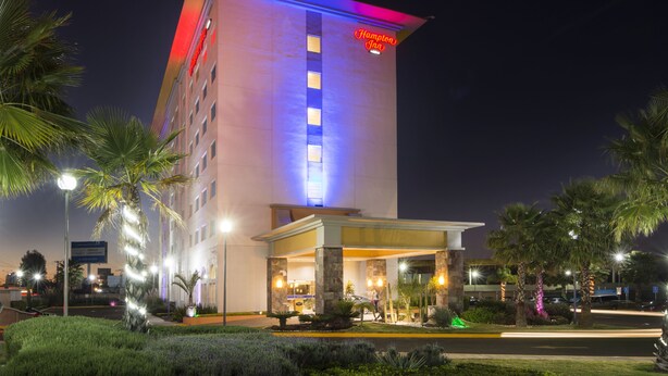 A hotel with a prominent entrance, palm trees, and a well-lit facade. - Hampton Inn by Hilton Silao-Aeropuerto Bajio (Silao)