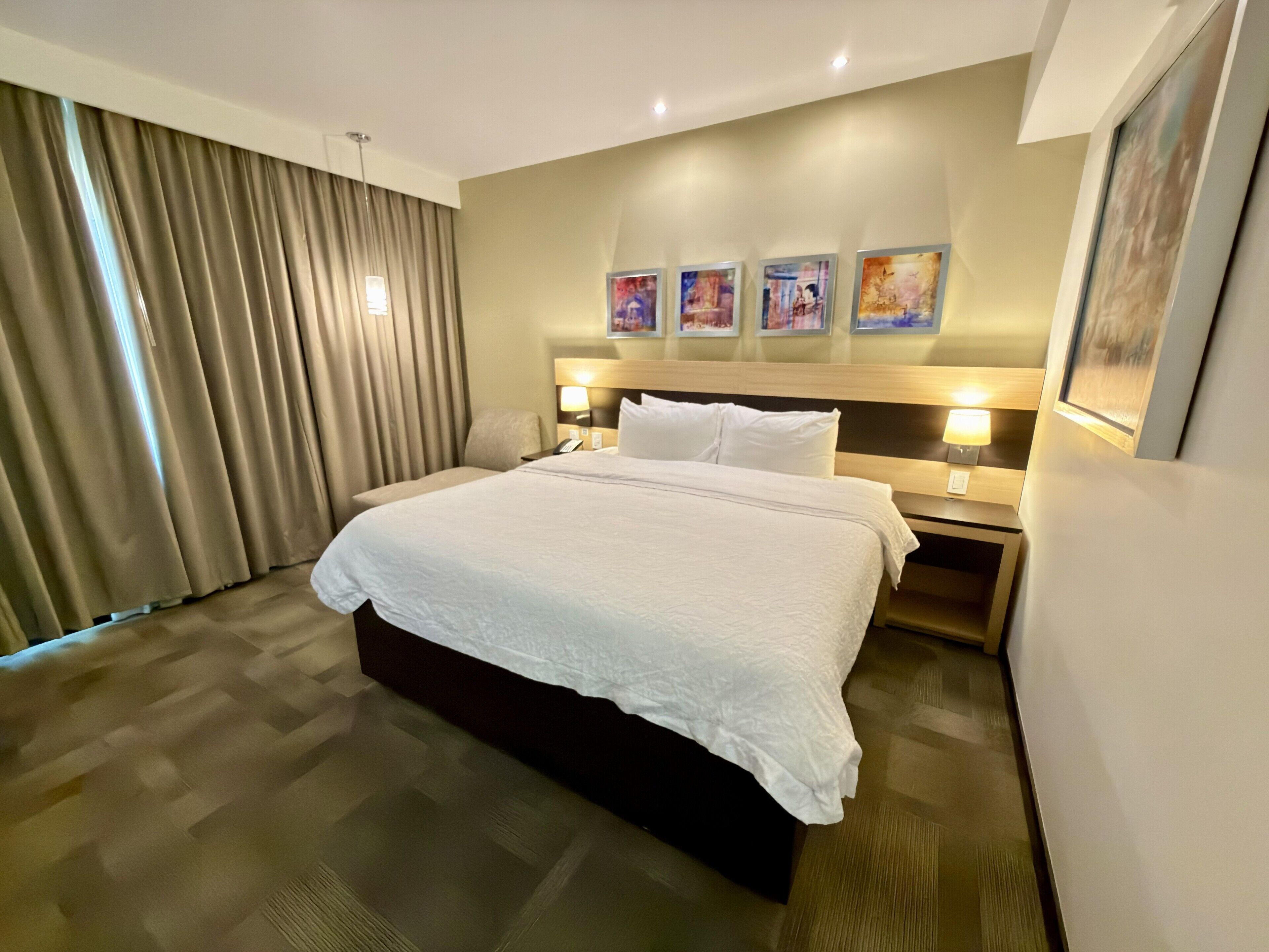 Quarto de hotel com uma cama grande, cabeceira com arte, mesa de cabeceira com abajur e janela com cortinas.