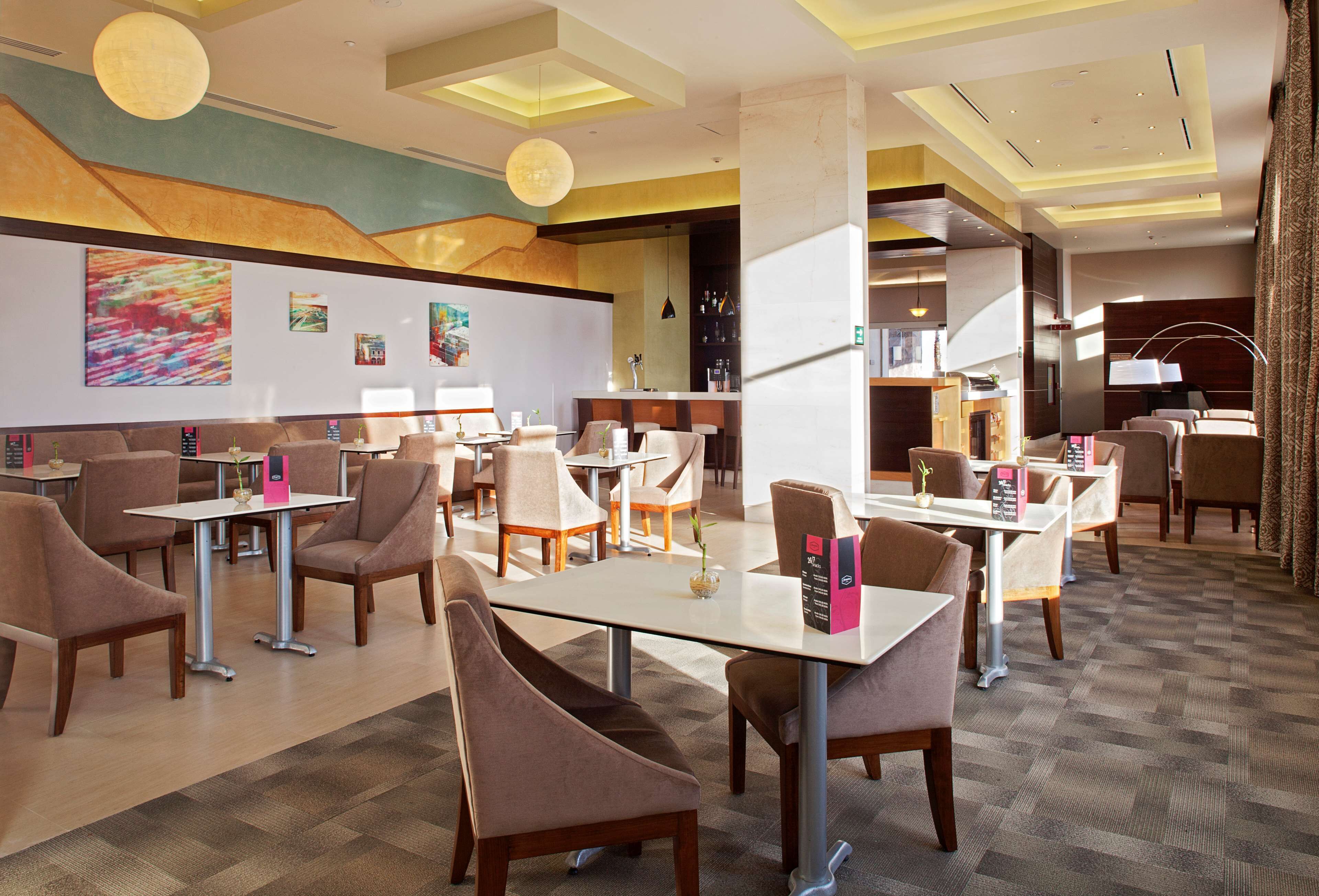 Un hall d’hôtel doté de plusieurs espaces aménagés avec des sièges, notamment des tables et des chaises, ainsi qu’un bar avec un comptoir et des tabourets.
