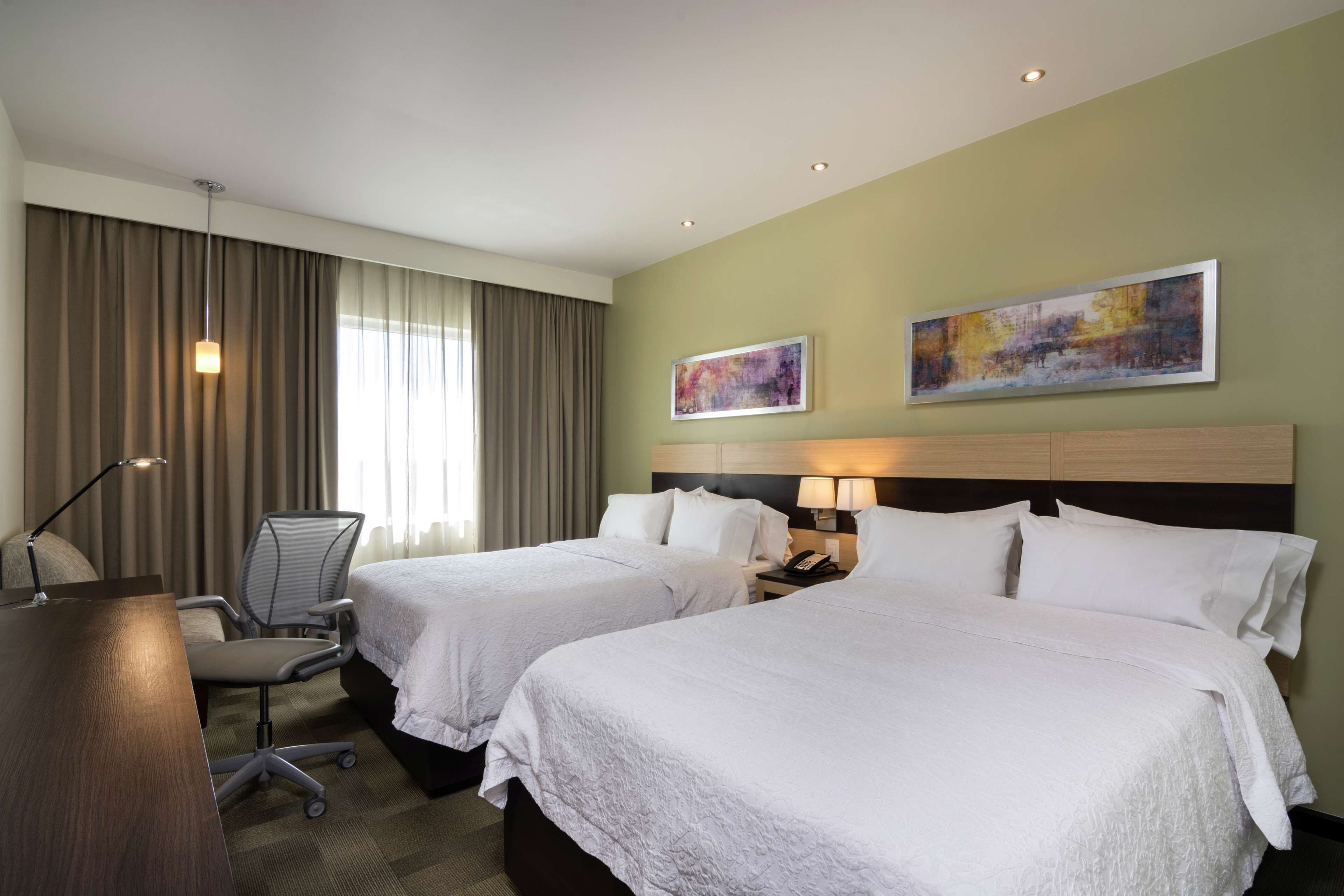 A hotel room with two beds, a desk, a chair, a lamp, and artwork on the walls.