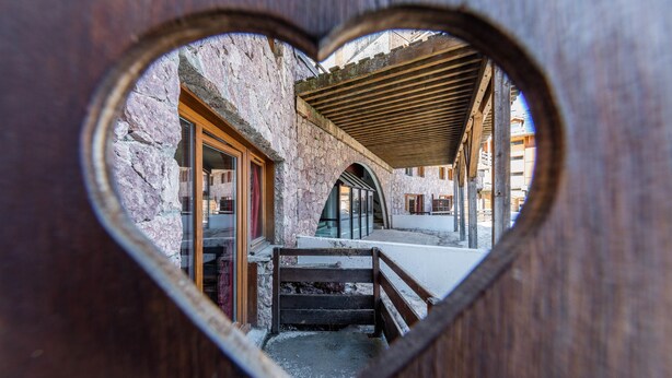 A heart-shaped window revealing a stone building with wooden beams and a covered walkway. - Noemys Castor & Pollux (Risoul)