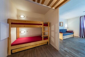 Un lit superposé avec un matelas rouge, un plancher en bois, une petite table avec un coussin et un luminaire fixé au mur.