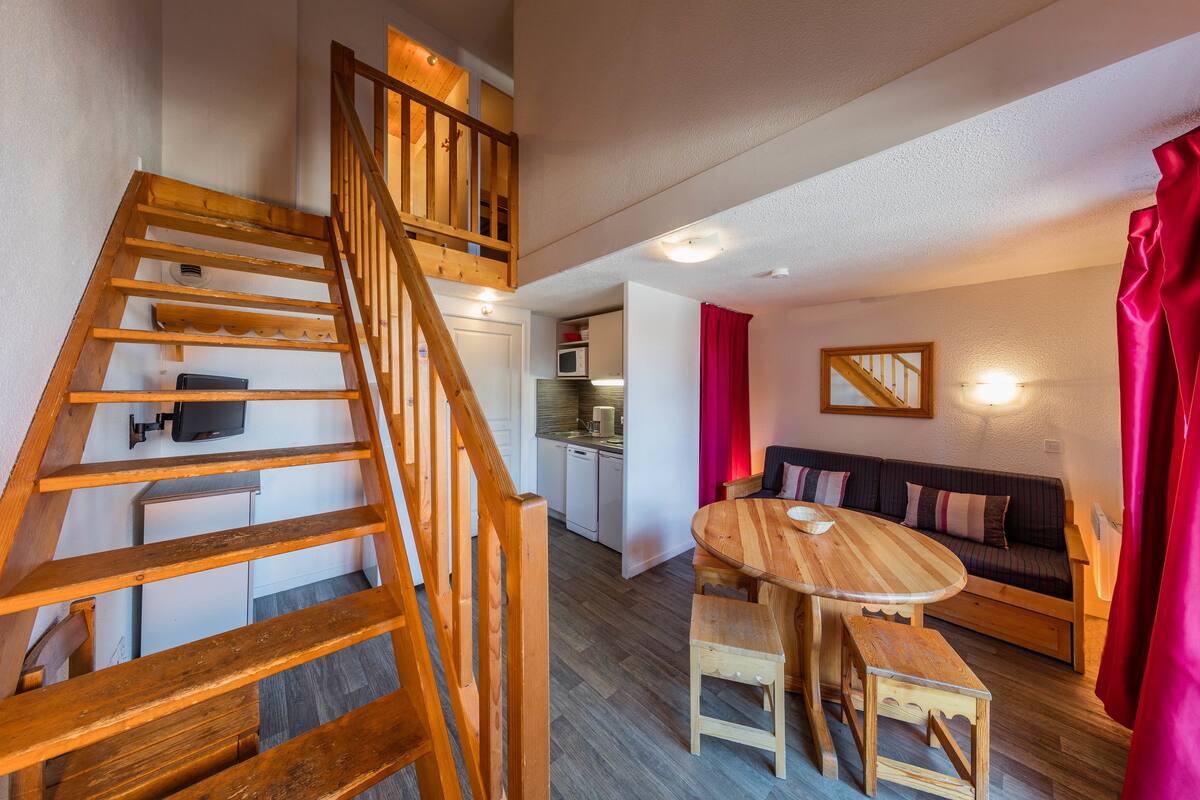 A wooden staircase leading to a loft bedroom, a dining area with a wooden table and bench, a kitchen with white appliances, and a living area with a sofa and red curtains.
