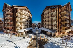 Un espace extérieur enneigé, avec des bâtiments en bois, un chemin central et un lampadaire.