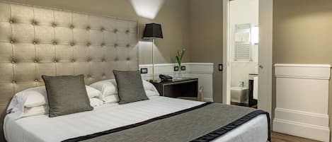 A neatly made bed with a tufted headboard, a bedside table with a lamp, and a bathroom visible through an open door.
