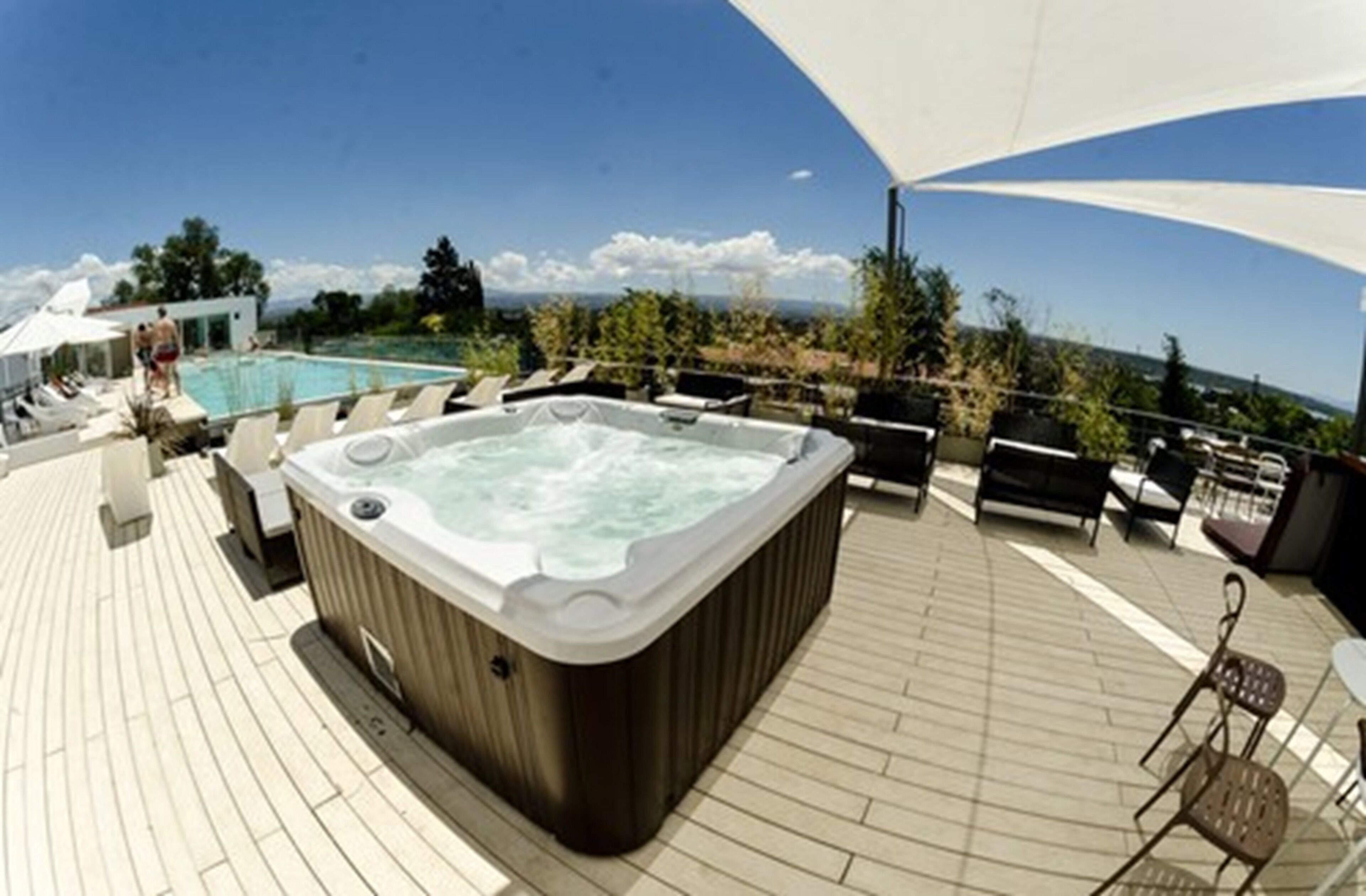 A hot tub on a wooden deck with a pool in the background and a clear sky.