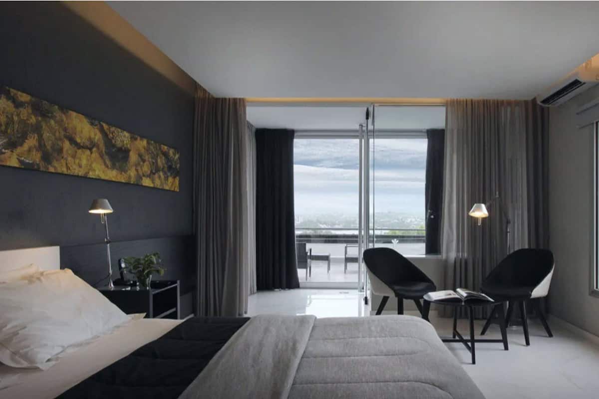 Une chambre d’hôtel moderne dotée d’un grand lit, d’un bureau, de deux chaises et offrant une vue sur la ville grâce à de grandes fenêtres.