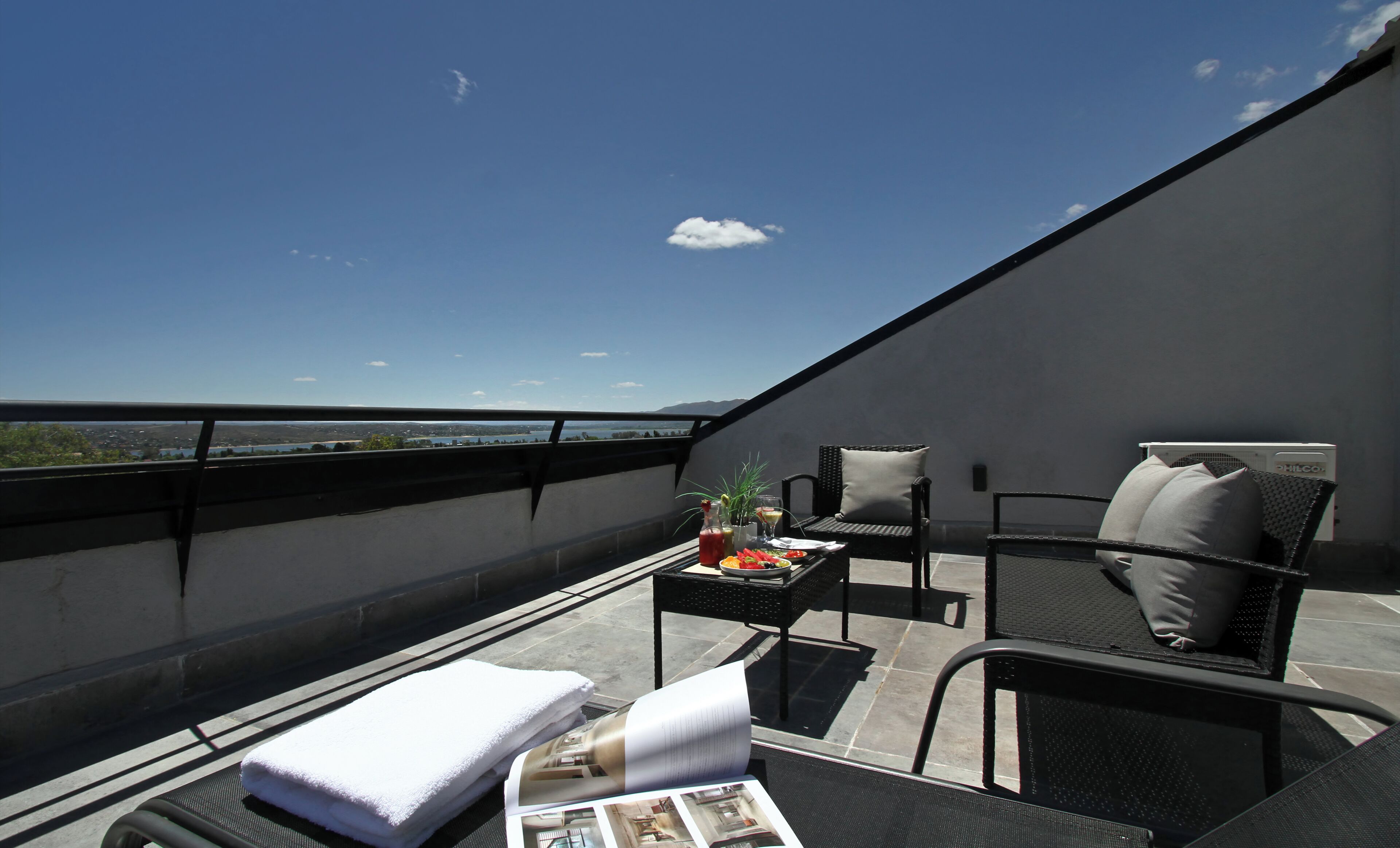 A rooftop terrace with a view of a lake and mountains, furnished with a table, chairs, and a towel.