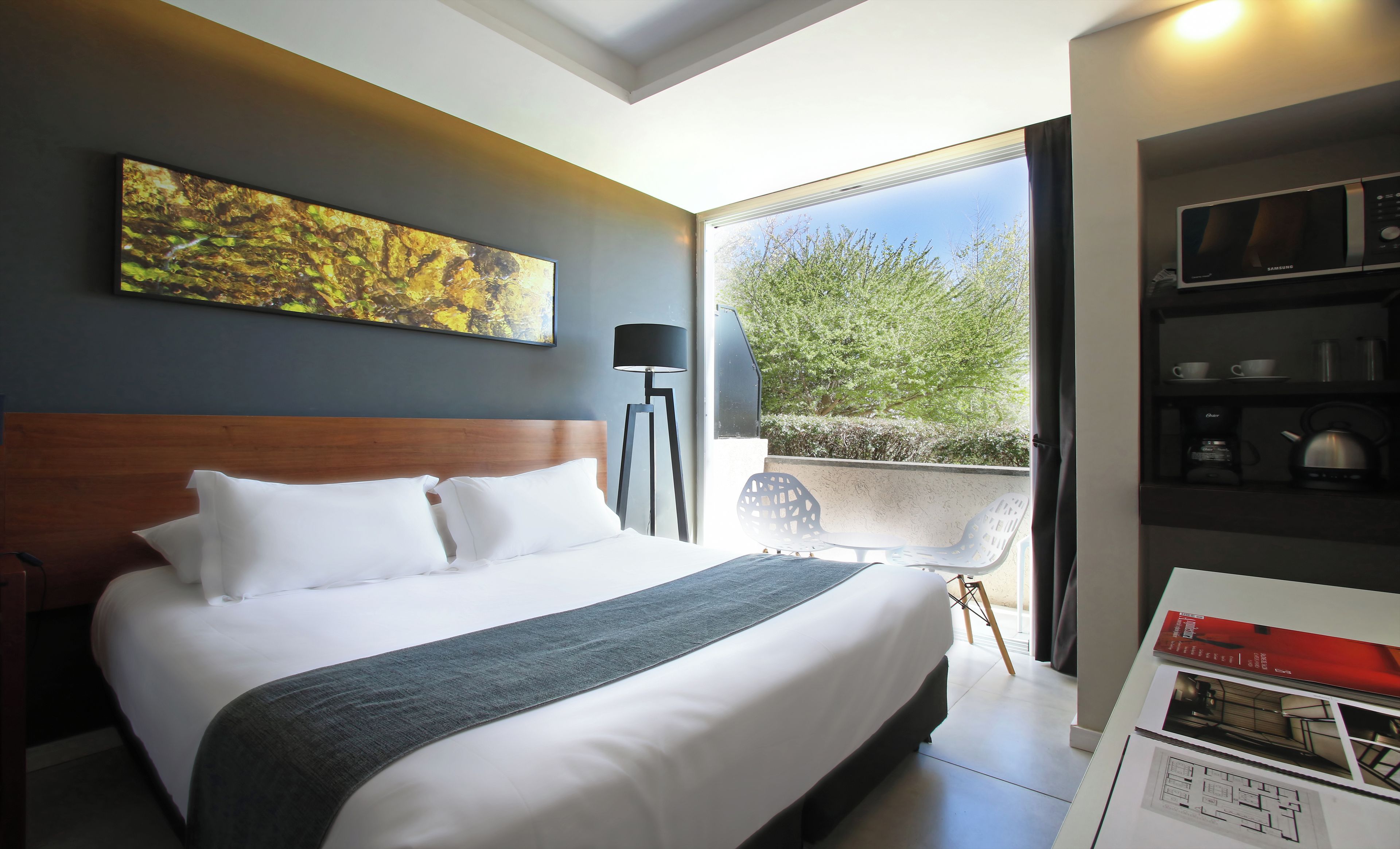 Habitación de hotel con una cama grande, vistas a zonas verdes, una lámpara de pie y una estantería con objetos.