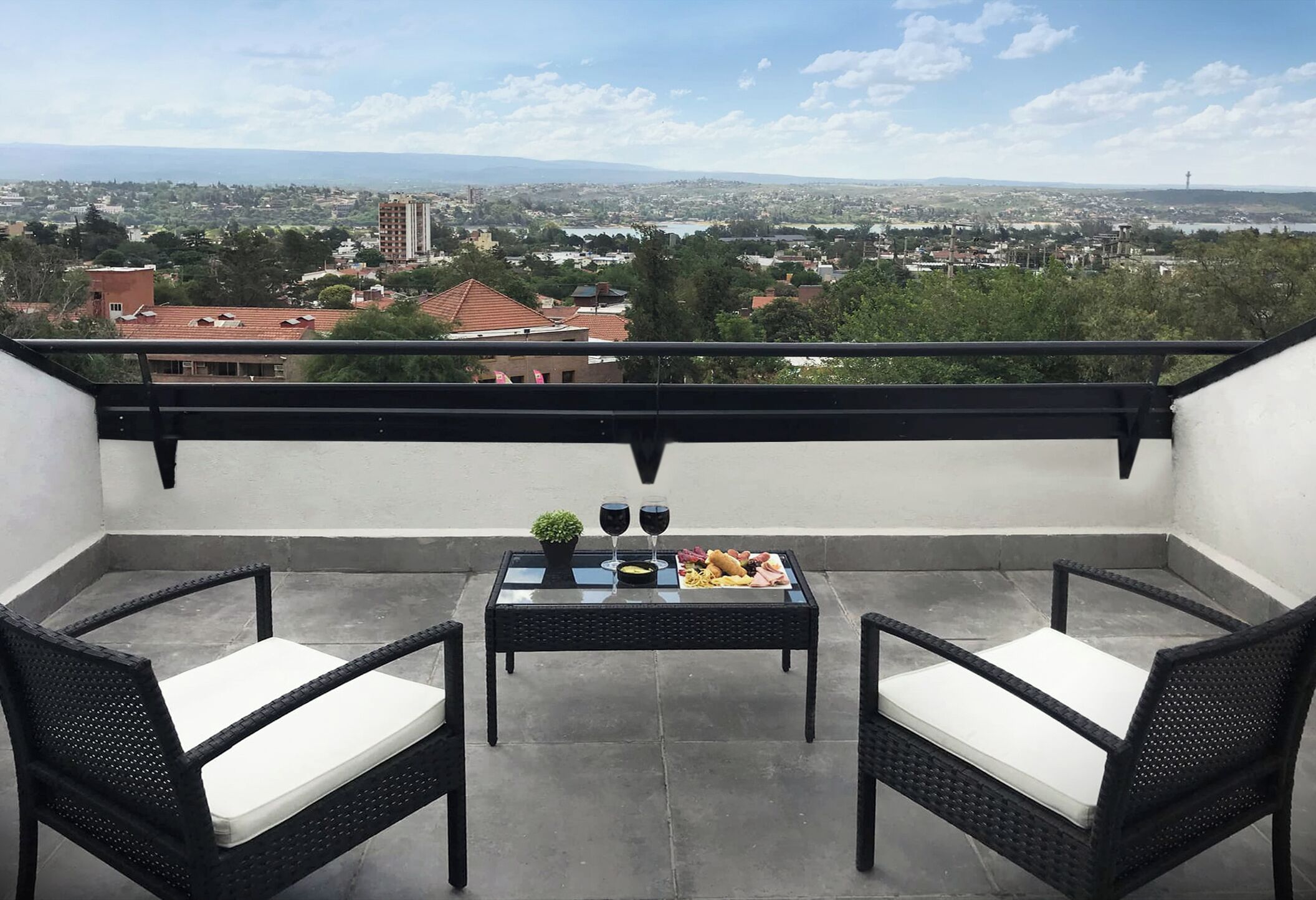 Un balcón con dos sillas de mimbre y una mesa dispuesta con comida y bebidas, con vista a un paisaje urbano.