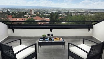 Un balcón con dos sillas de mimbre y una mesa dispuesta con comida y bebidas, con vista a un paisaje urbano.