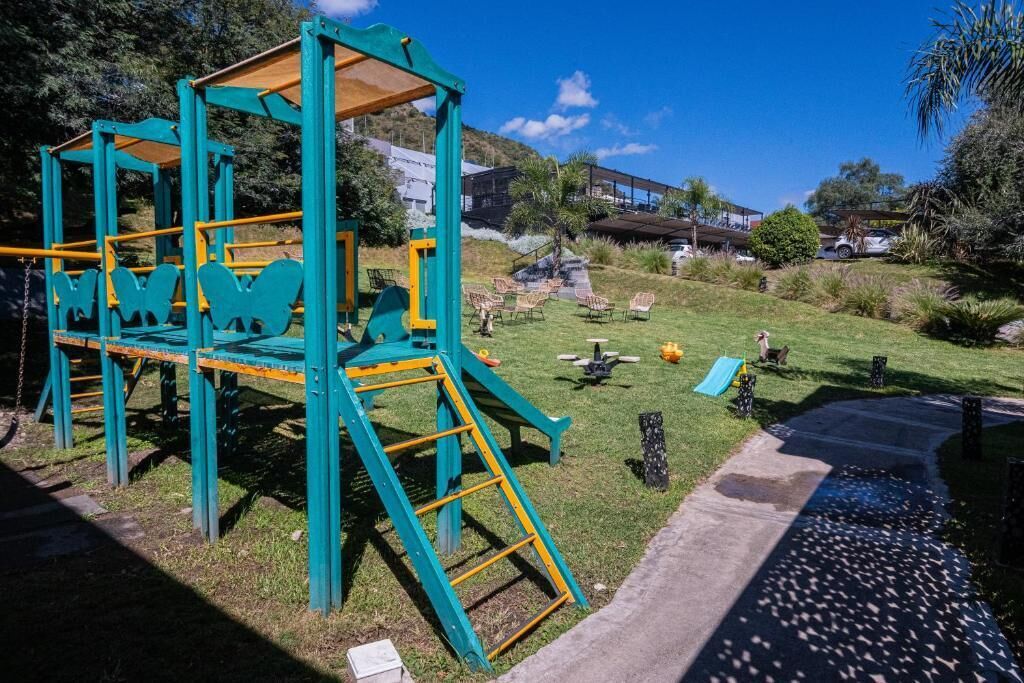 Un área de juegos con estructuras para escalar y toboganes, rodeada de césped, sillas y un sendero de concreto.