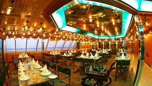 Un comedor con mesas dispuestas para una comida, ventanas amplias y un alto techo con lámparas colgantes.