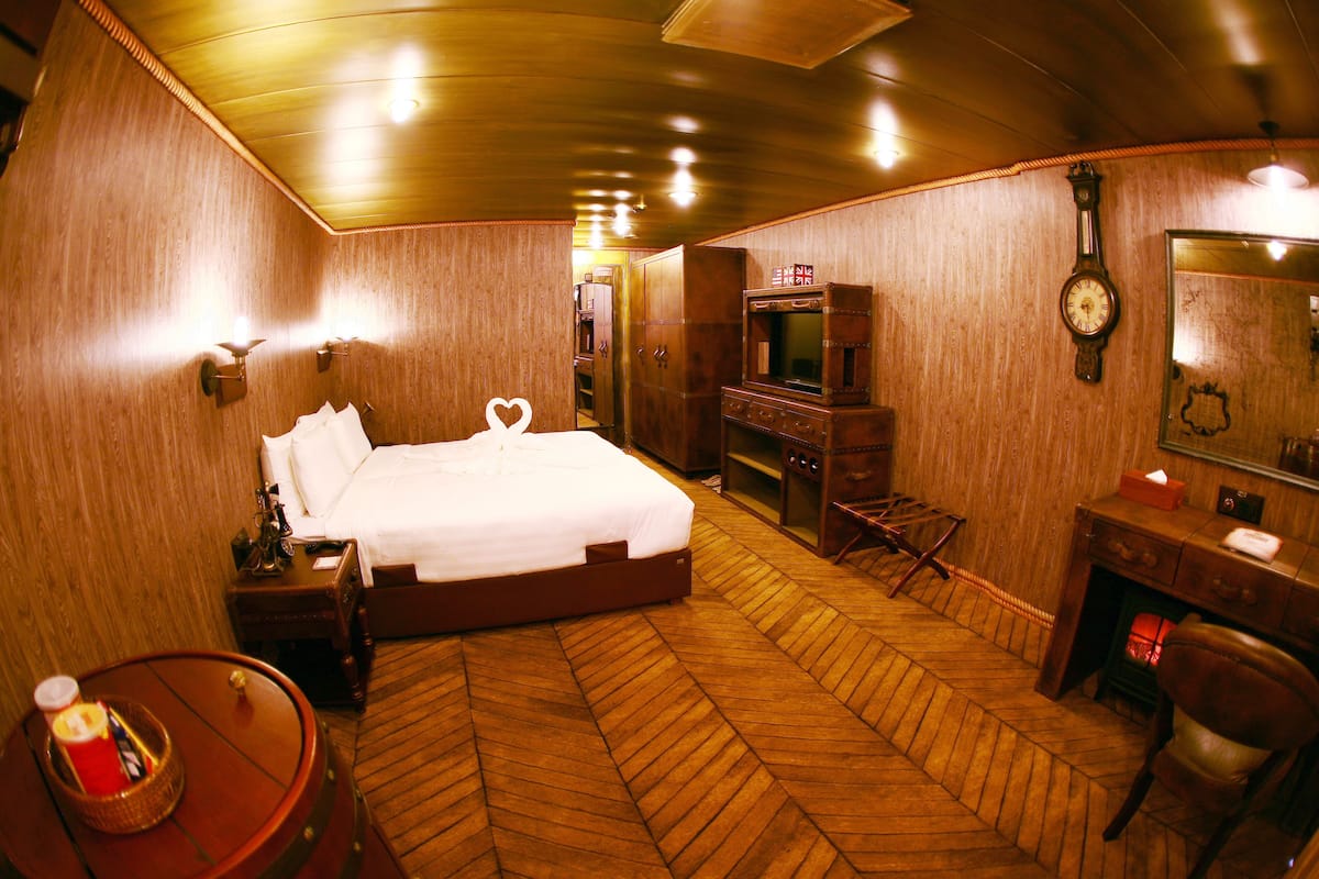 Une chambre d’hôtel avec un parquet en bois disposé en chevrons, un lit avec du linge de lit blanc, une commode en bois, un bureau avec une chaise, une horloge au mur et un miroir au-dessus de la commode.