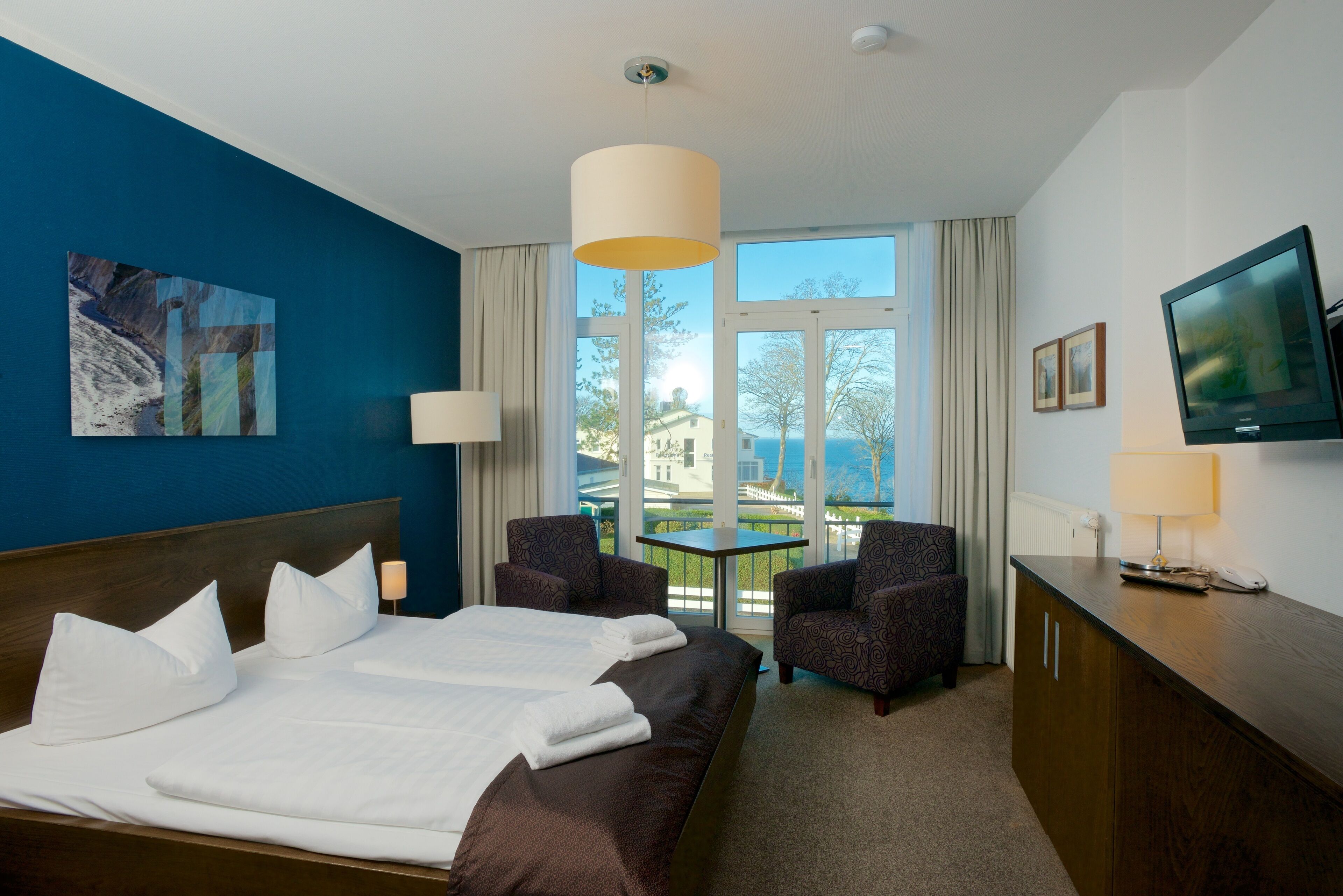 A hotel room with a large bed, two armchairs, a small table, a TV, and a window with a view of buildings and trees.