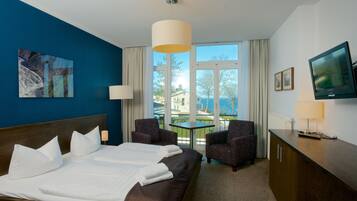 A hotel room with a large bed, two armchairs, a small table, a TV, and a window with a view of buildings and trees.