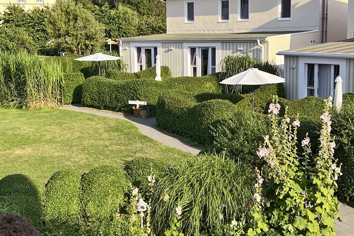 Un jardin bien entretenu, avec des haies taillées avec soin et un chemin menant à un bâtiment doté de nombreuses fenêtres et agrémenté de parasols d’extérieur.