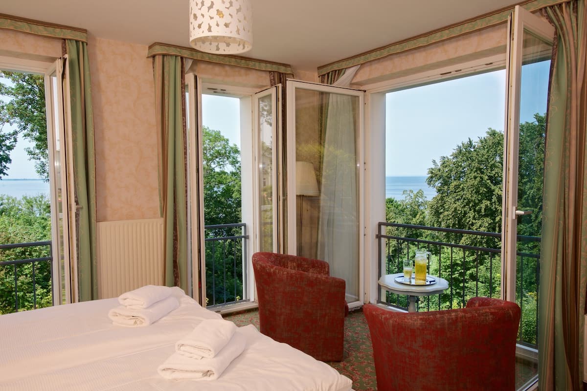 Une chambre d’hôtel avec un lit, deux fauteuils rouges, une petite table avec une boisson, et une vue sur la mer par de grandes fenêtres.