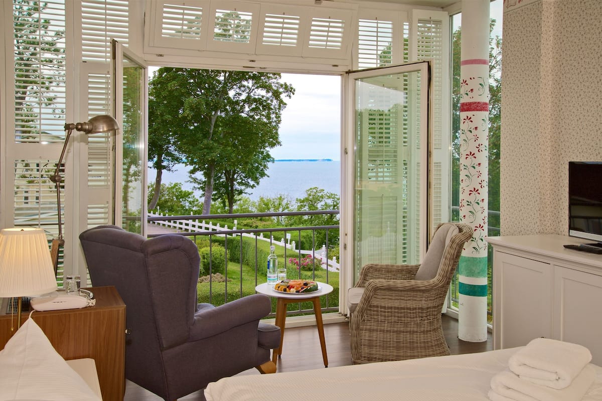 Un balcon avec vue sur la mer, une chaise en osier, une petite table avec une assiette de nourriture et un téléviseur posé sur un meuble.