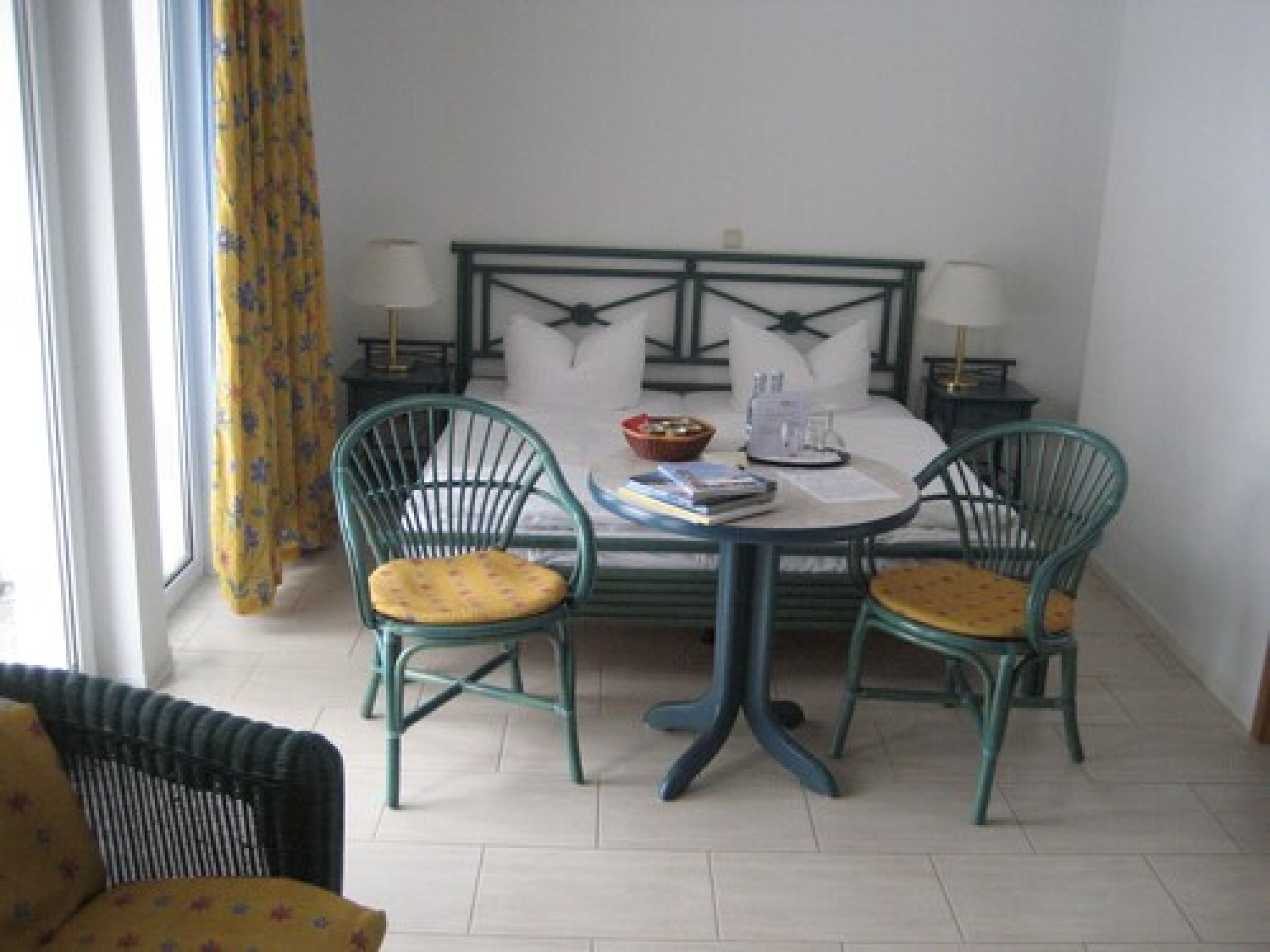 Un coin repas avec une table ronde, deux chaises, un lit avec une tête de lit et une fenêtre avec des rideaux.