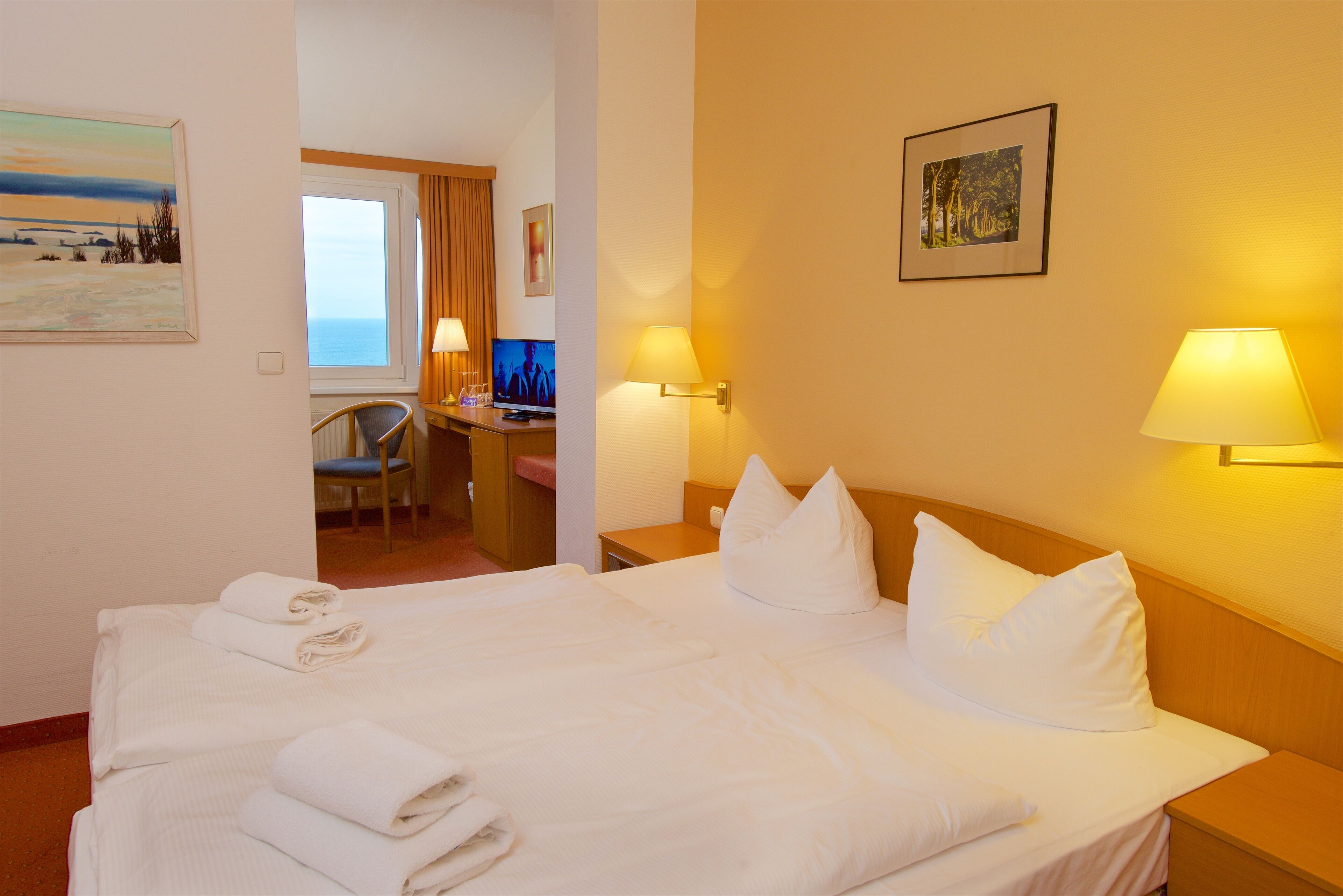 A hotel room with a large bed, two bedside lamps, a desk with a computer, and a chair.