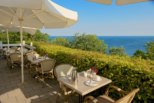 An outdoor dining area with tables and chairs, umbrellas, and a view of the sea.