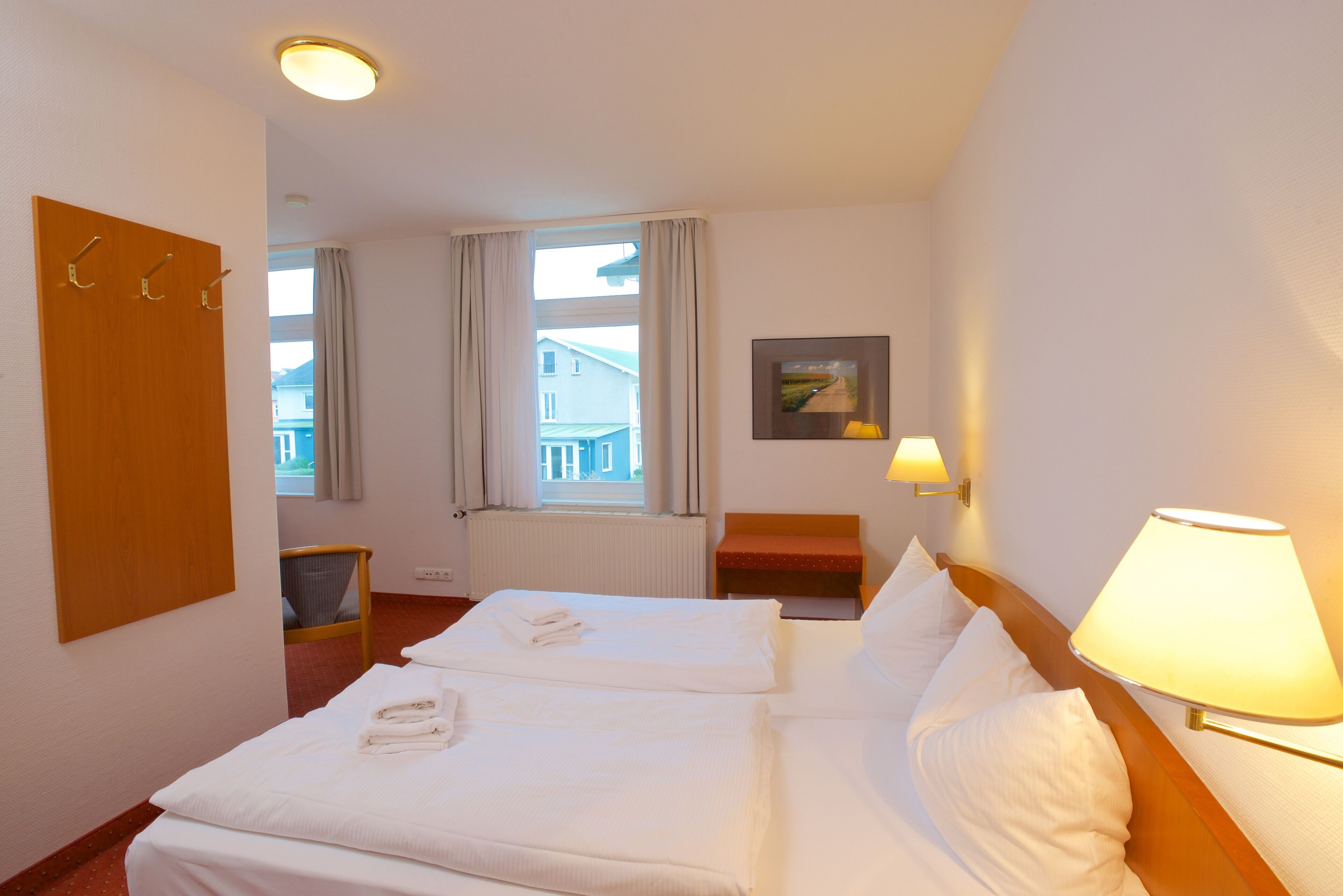 Une chambre d’hôtel avec deux lits, une porte en bois avec des crochets, une fenêtre avec des rideaux, une chaise, une petite table, une lampe fixée au mur et un tableau encadré.