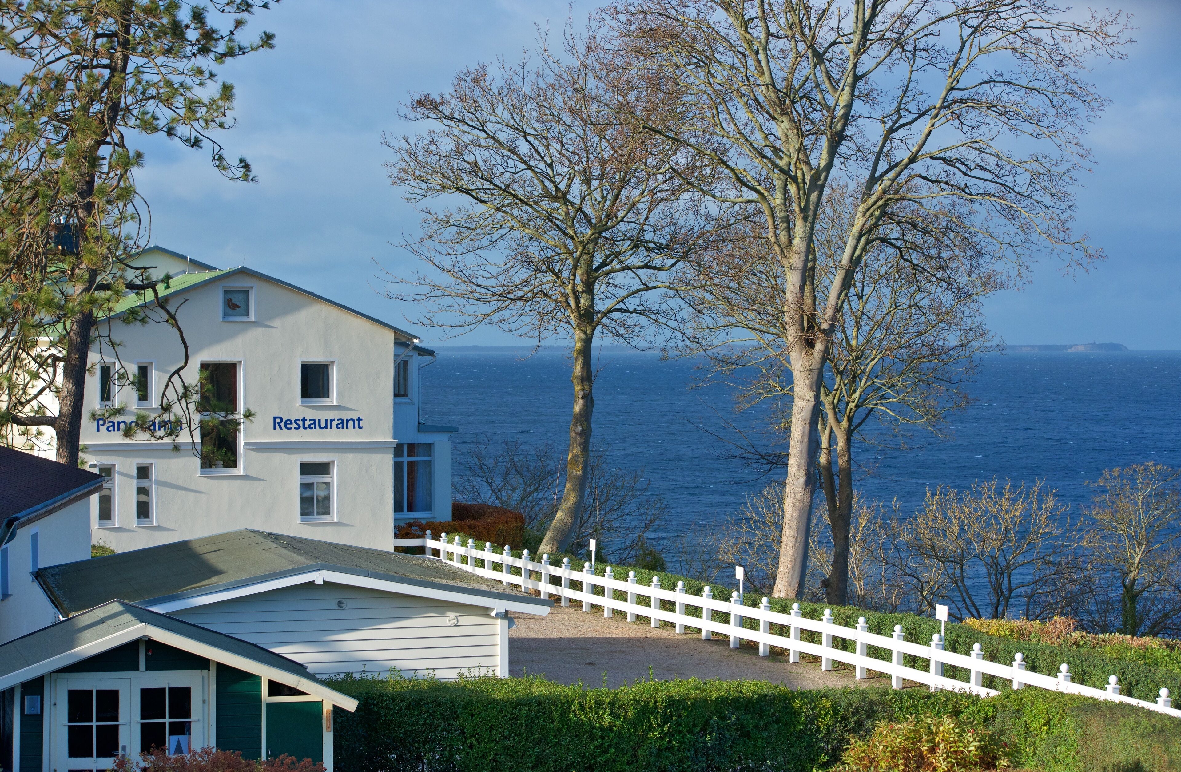 A restaurant with a view of the sea, a white fence, and a building with a green roof.