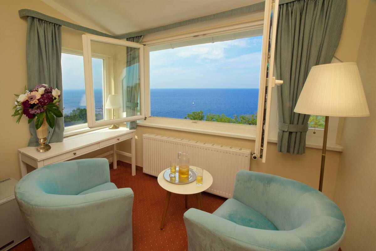Une pièce avec deux fauteuils bleu clair, une petite table sur laquelle se trouvent une carafe et un verre, un miroir, un radiateur et une vue sur la mer.