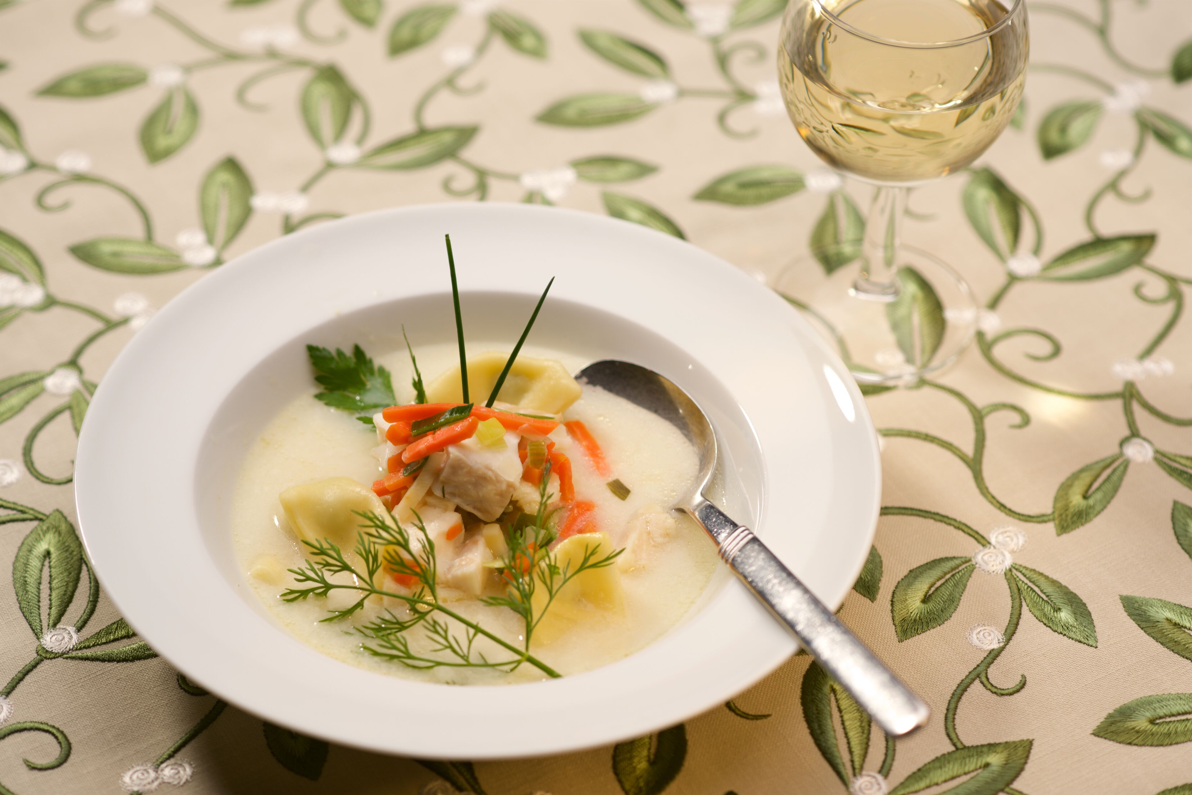 A bowl of soup with vegetables and a spoon, placed on a table with a floral tablecloth.