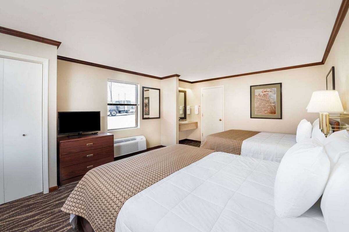 A hotel room with two beds, a TV, a dresser, a window, a mirror, and a framed picture on the wall.