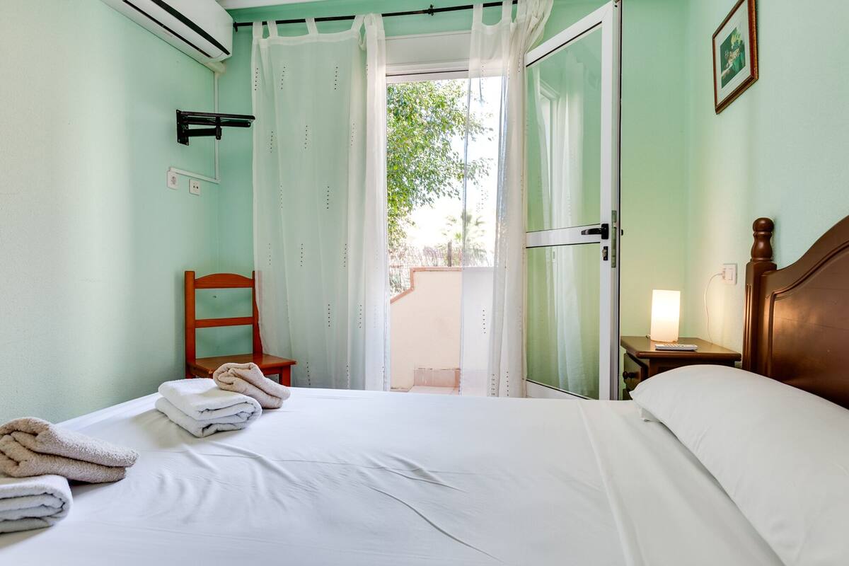 A hotel room with a bed, a chair, a bedside table with a lamp, and a view of the outside.