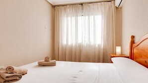 A hotel room with a neatly made bed, a wooden headboard, a bedside table with a lamp, and a window with sheer curtains. - ApartHotel Atlas (Torrevieja)