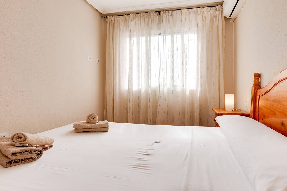 A hotel room with a neatly made bed, a wooden headboard, a bedside table with a lamp, and a window with sheer curtains.