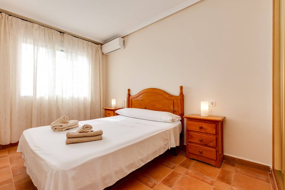 A neatly made bed with a wooden headboard, a bedside table with a lamp, and a window with sheer curtains.