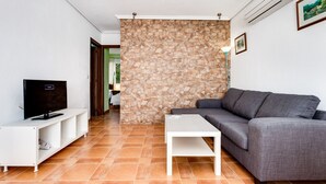 A living room with a stone wall, a grey sofa, a white coffee table, and a TV on a white stand. - ApartHotel Atlas (Torrevieja)
