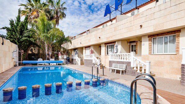 A swimming pool with a tiled deck, surrounded by palm trees and a building with a sign reading "AP". - ApartHotel Atlas (Torrevieja)