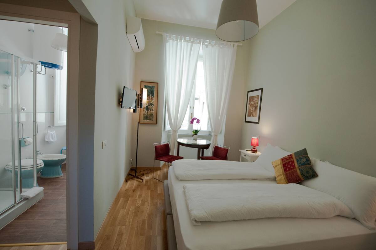 Una habitación de hotel con cama, escritorio con lámpara, silla, ventana con cortinas y baño con ducha e inodoro.