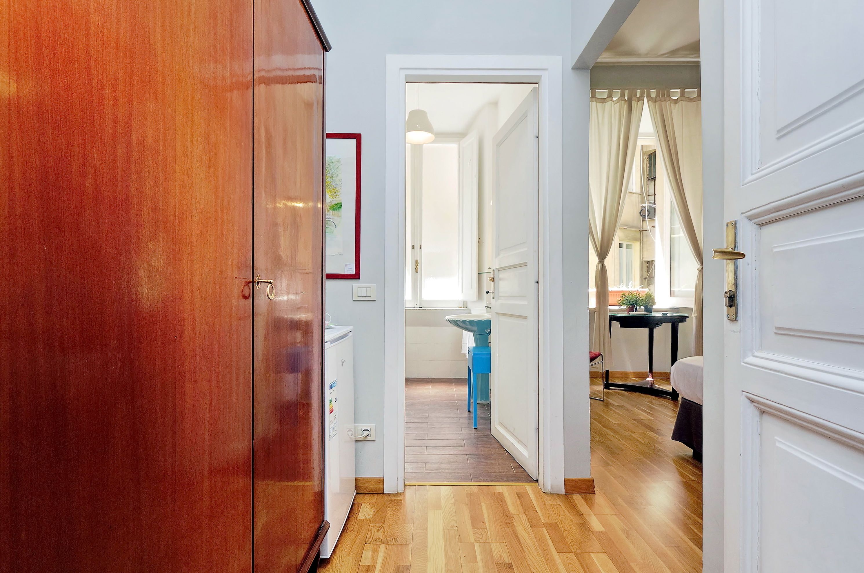 A hallway with a wooden wardrobe, a white door, and a view into a room with a bed, a table, and a window with curtains.