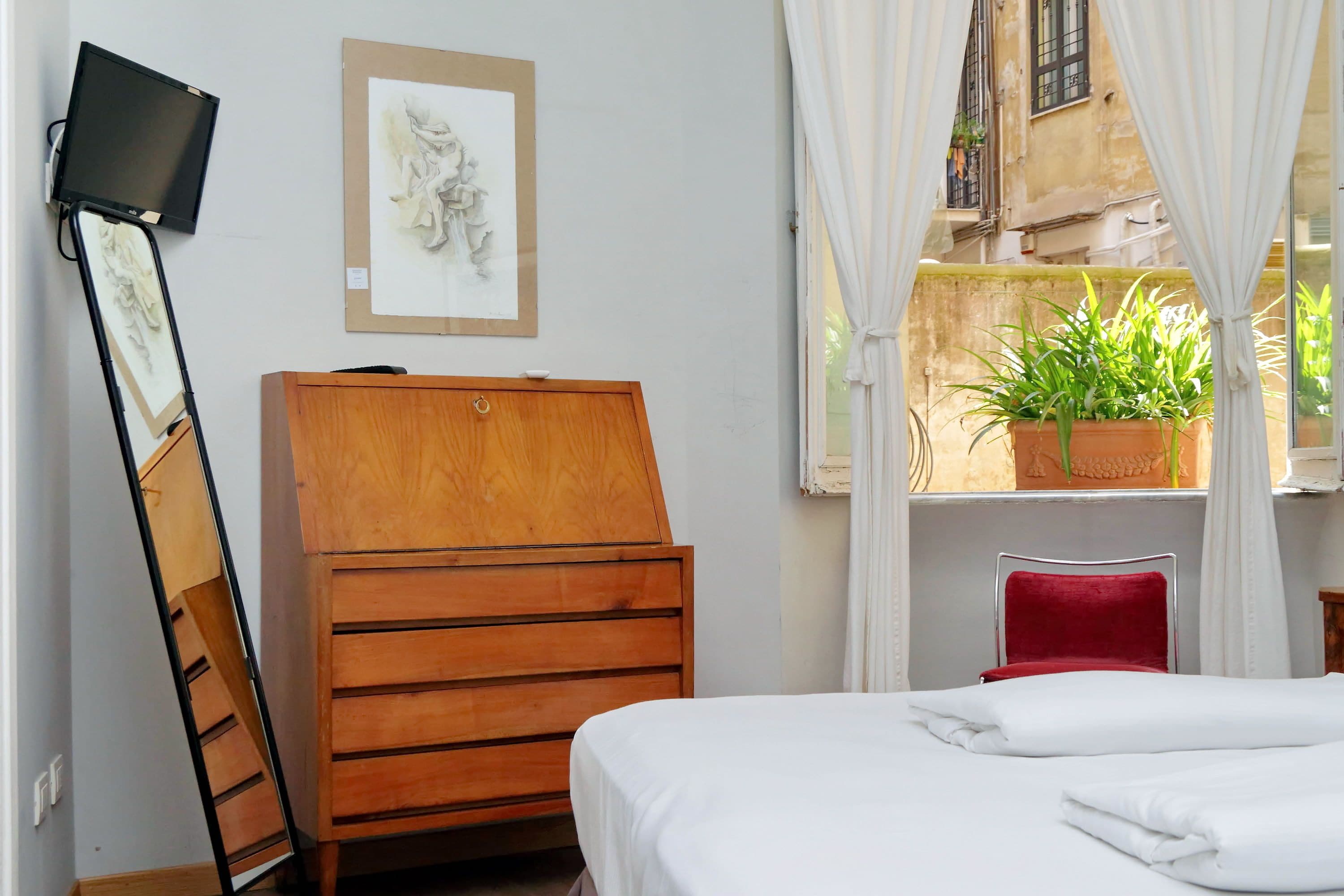 A bedroom with a bed, wooden dresser, a flat-screen TV mounted on the wall, a framed artwork, a window with curtains, and a potted plant.