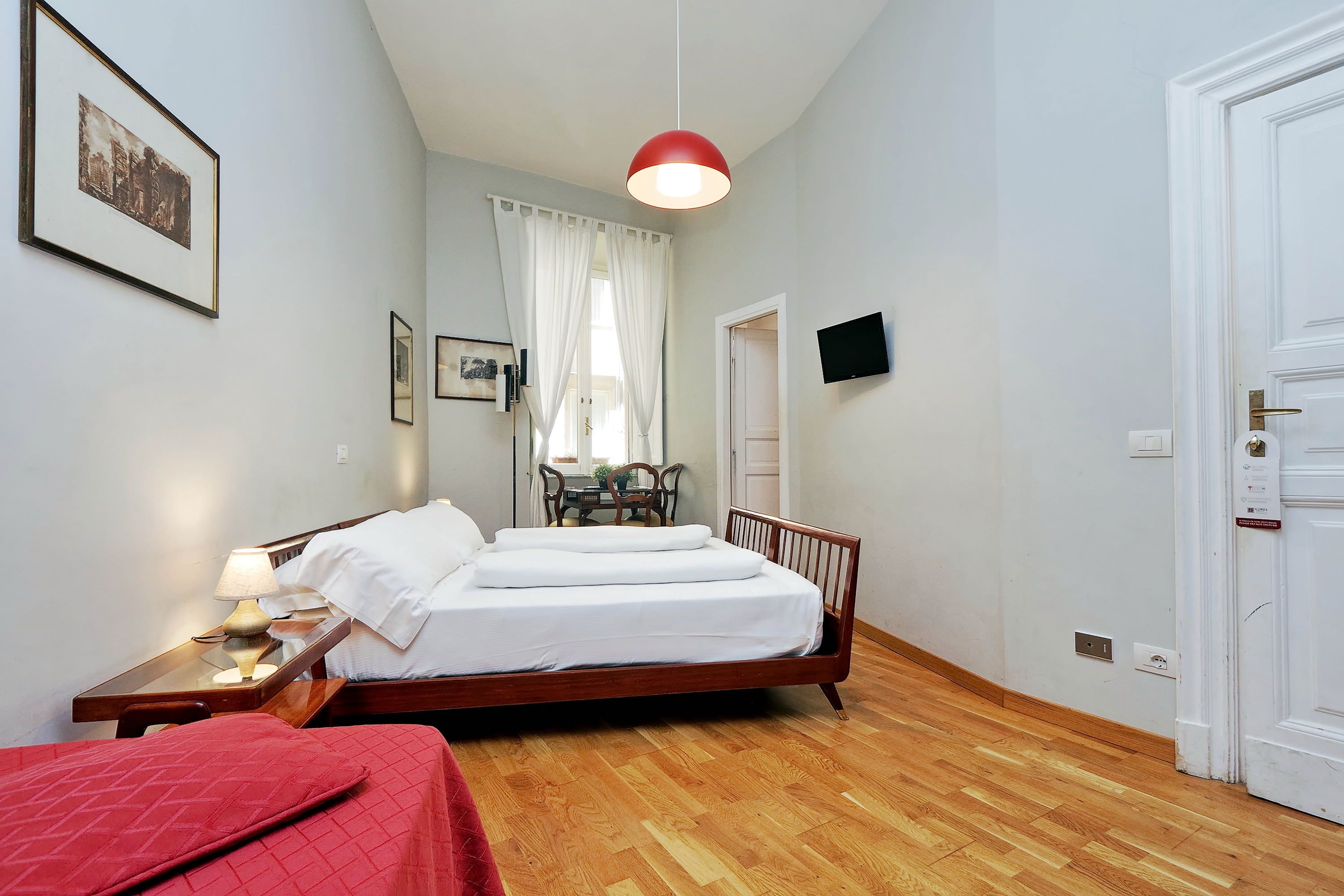 Un dormitorio con una cama de madera, una mesita de noche con lámpara, una colcha roja, una ventana con cortinas blancas y un televisor en la pared.