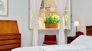 Quarto de hotel com cama, cómoda de madeira, cadeira vermelha, janela com cortinas brancas, planta em vaso e mesa de cabeceira com abajur.