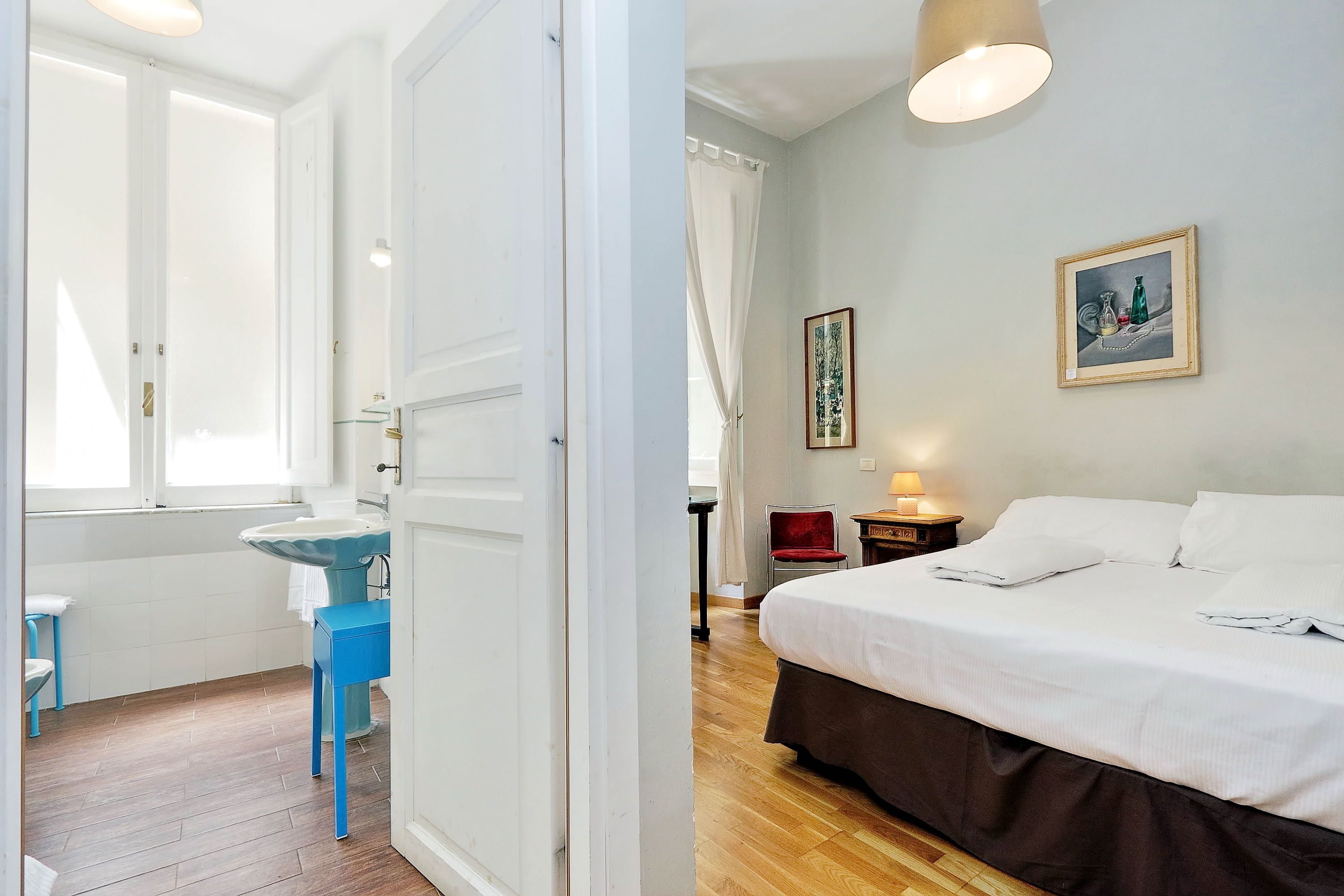 A hotel room with a bed, a bedside table, a chair, a window, and a bathroom with a sink and mirror.