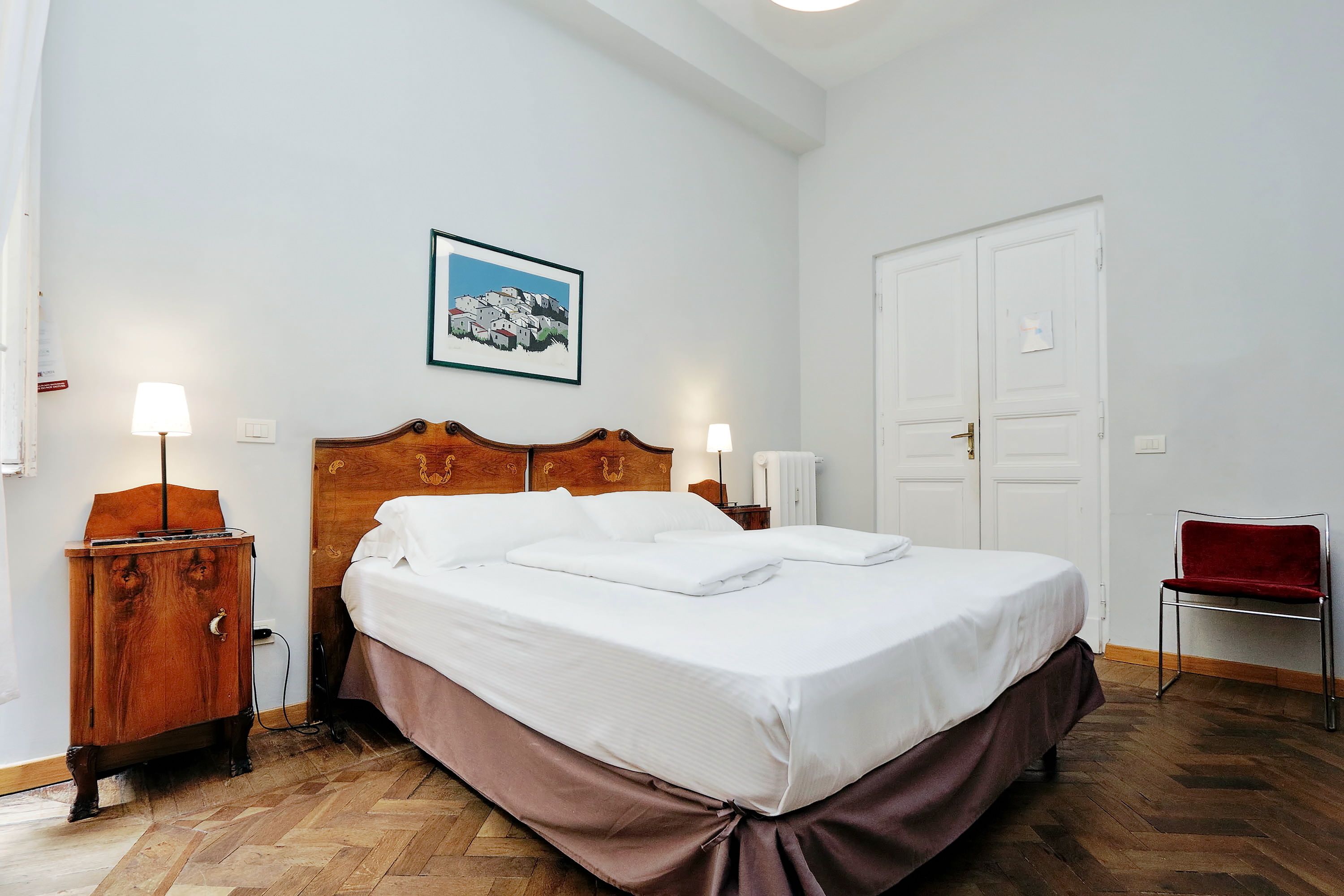 A neatly made bed with a wooden headboard, a wooden nightstand with a lamp, a red chair, and a white door.