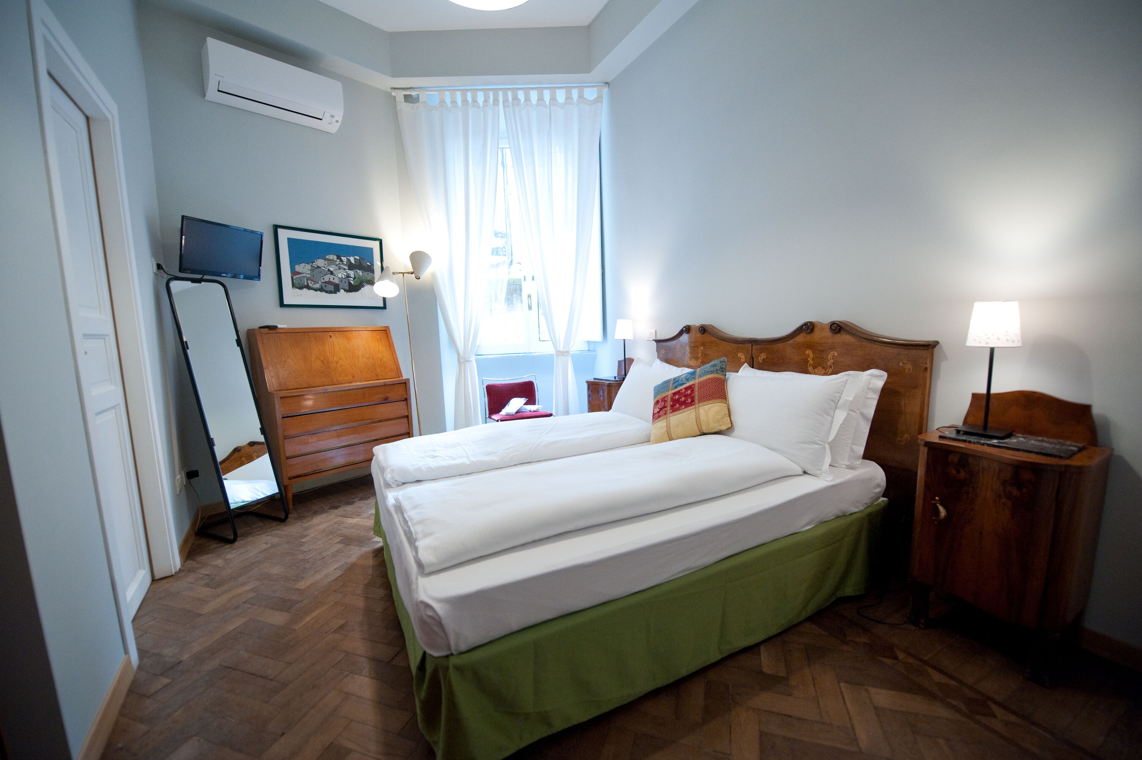 A hotel room with a wooden bed, a green skirt, a wooden nightstand, a mirror, a picture on the wall, and a window with curtains.